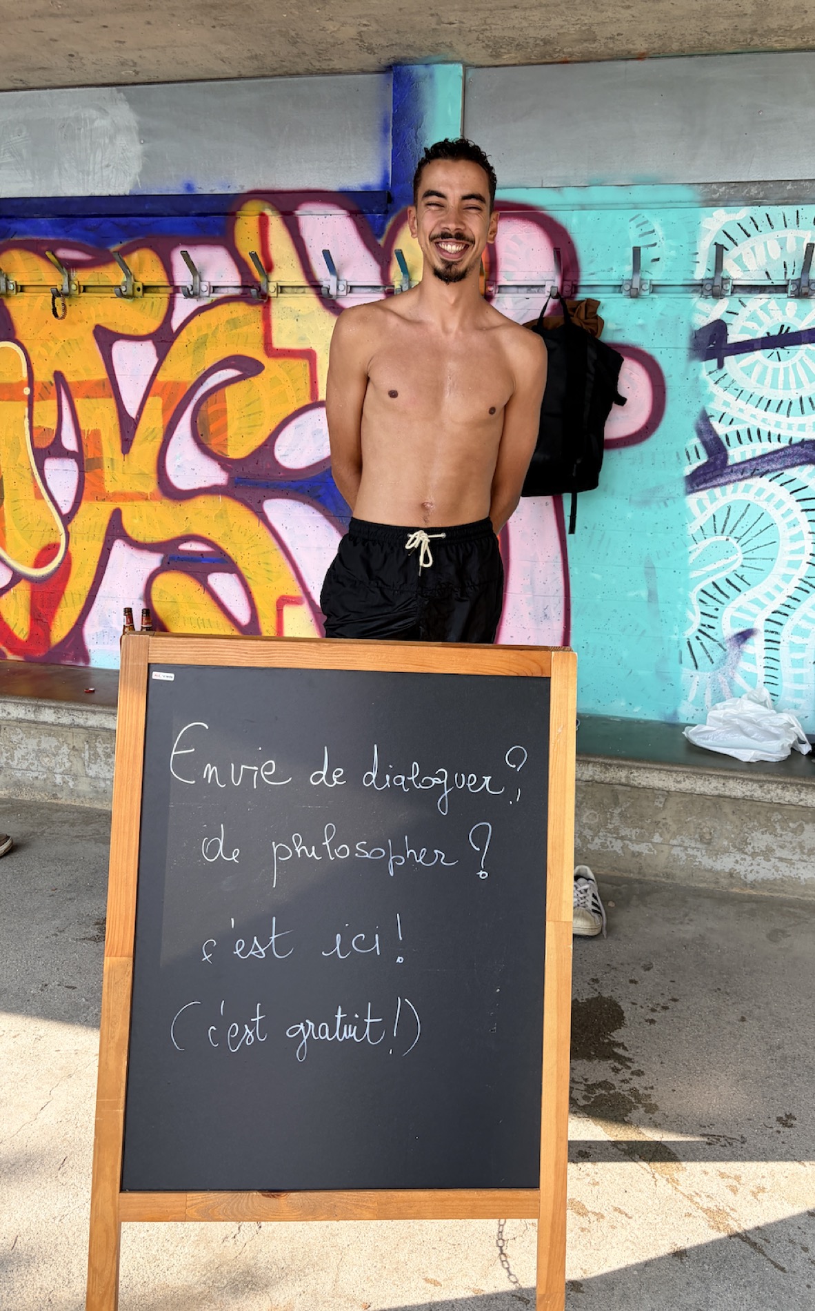 Un passant pose en riant derrière la pancarte, sous un mur couvert de graffitis colorés.