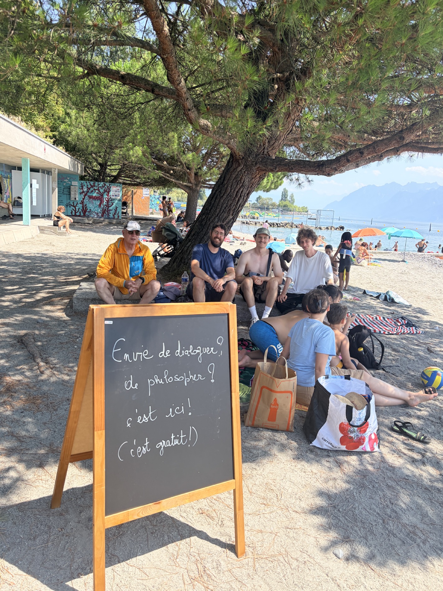 Un dialogue improvisé sous un grand pin, sur la plage de Vidy, avec le lac et les montagnes en arrière-plan.