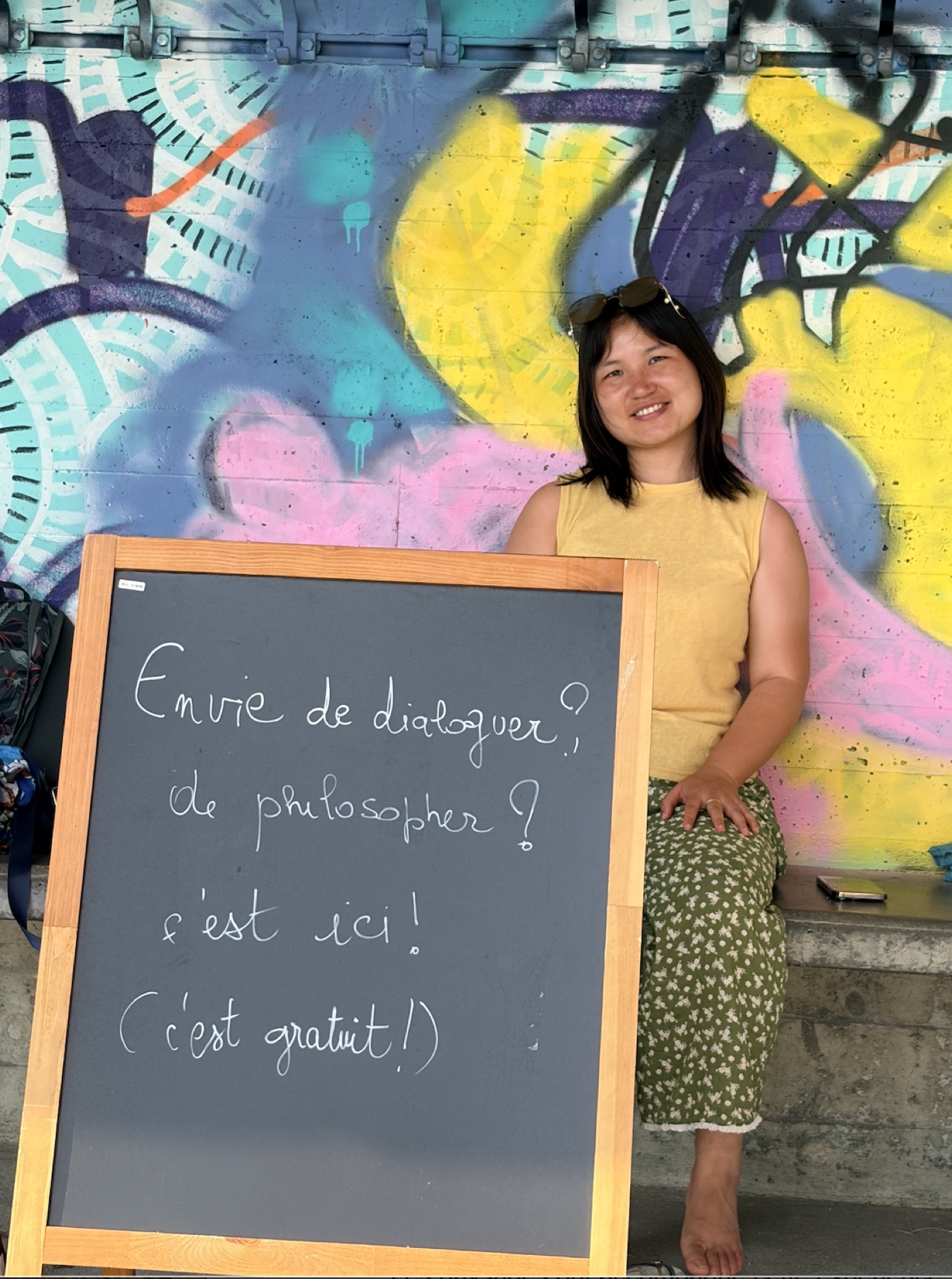 Portrait de Wei, souriante, assise près de la pancarte devant un mur de graffitis colorés.